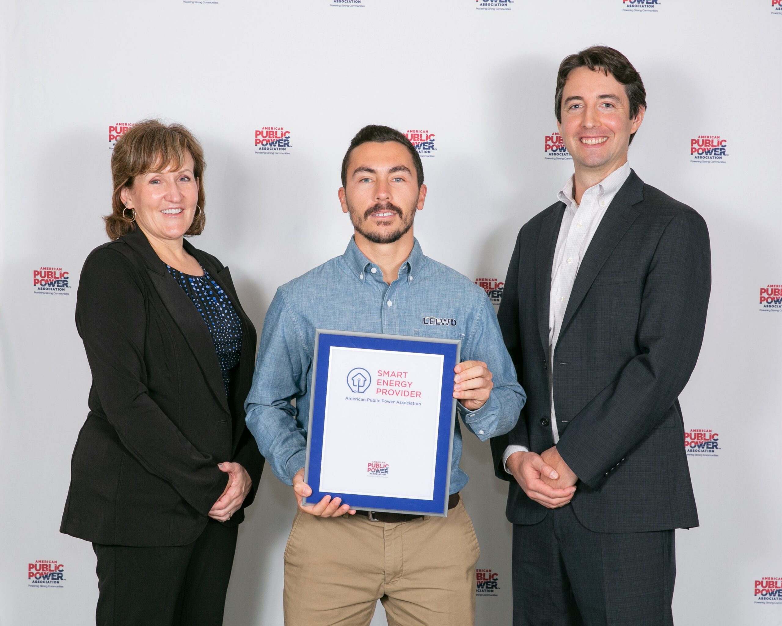 Three people pose for a photo at an awards event, center person holding a blue-framed certificate recognizing a Smart Energy Provider designation.