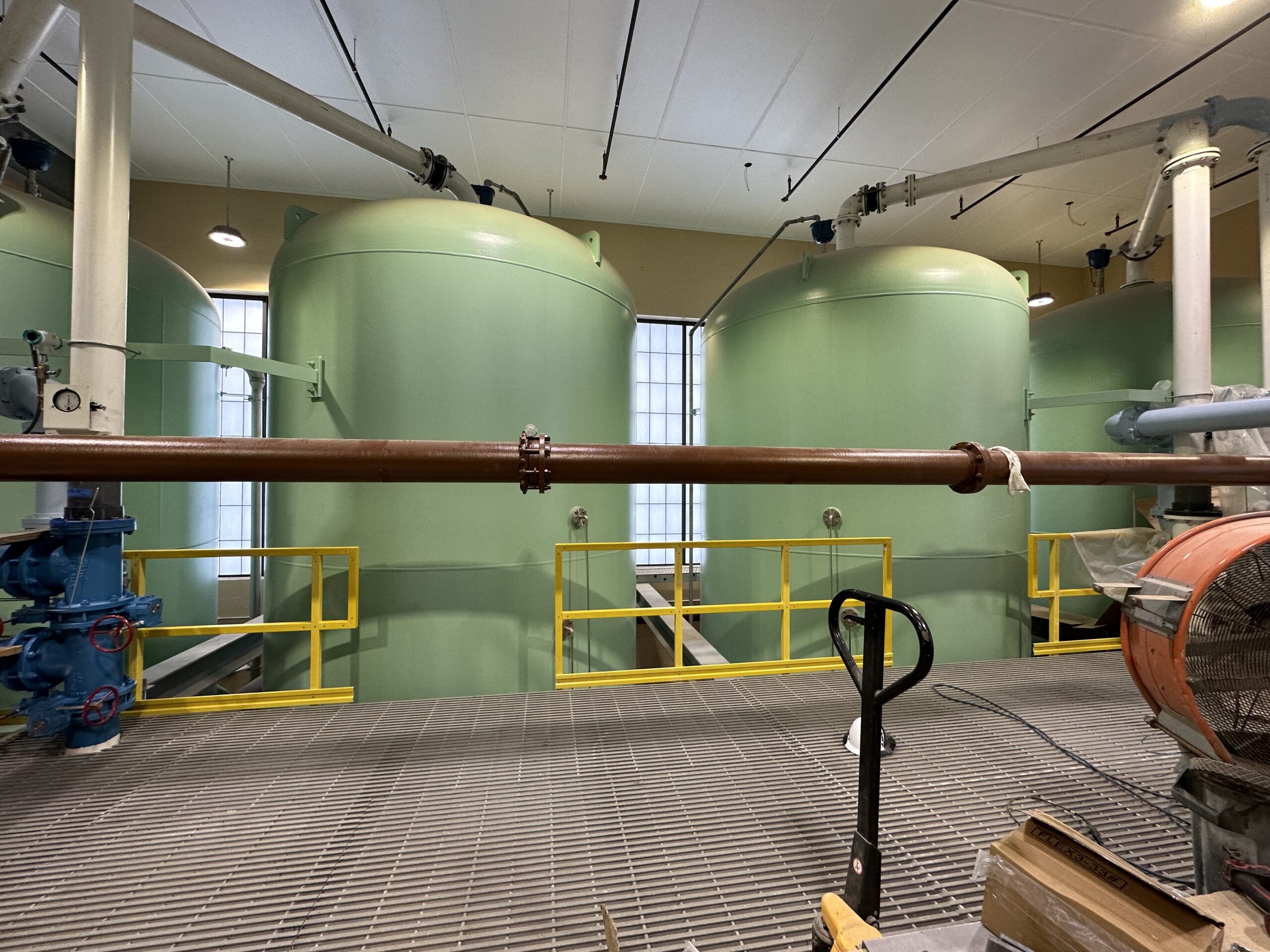 Indoor view of a water treatment plant featuring large green cylindrical tanks, yellow safety rails, and networked pipes above a metal grating floor.