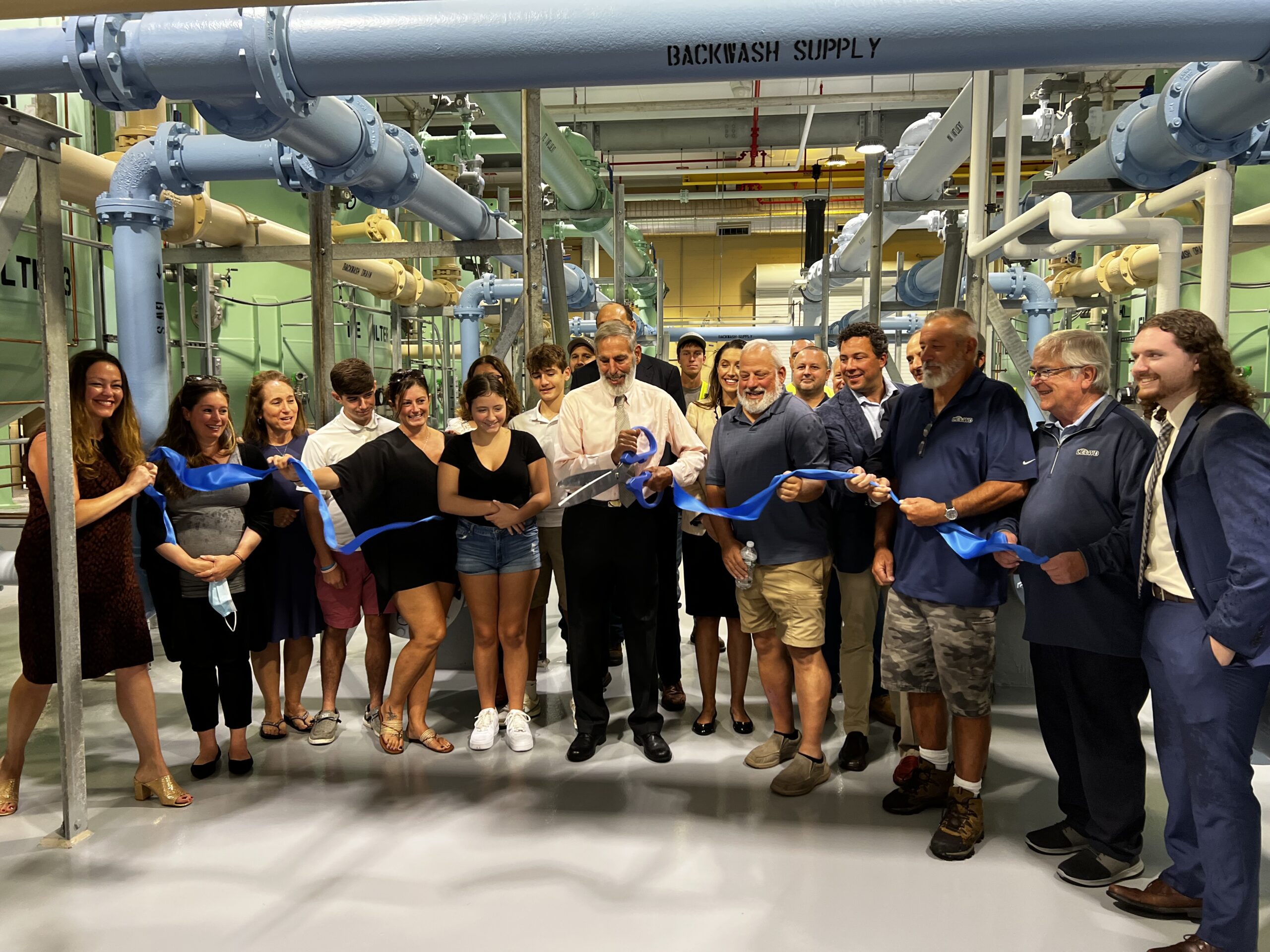 Officials and community members participate in a ribbon-cutting ceremony inside a water treatment plant, standing beneath large blue piping.