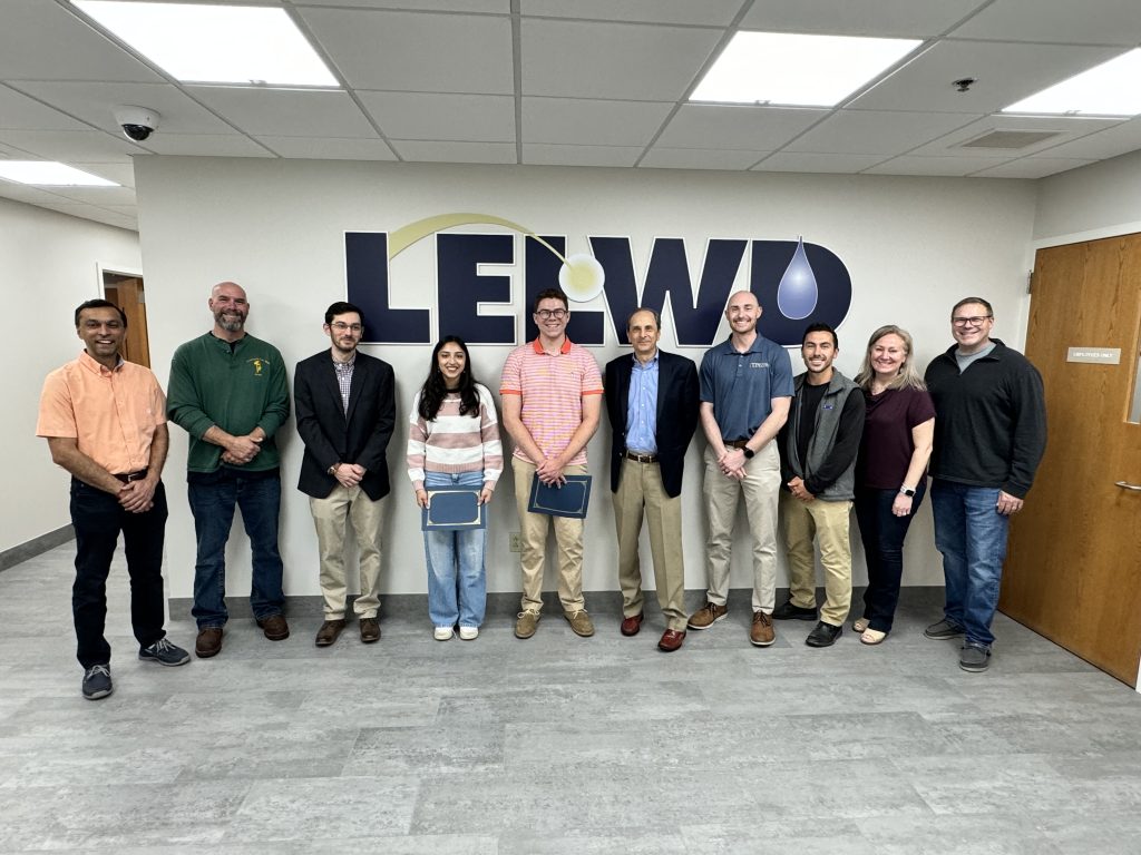 Eleven people pose for a group photo indoors in front of a wall bearing an LELWD logo.