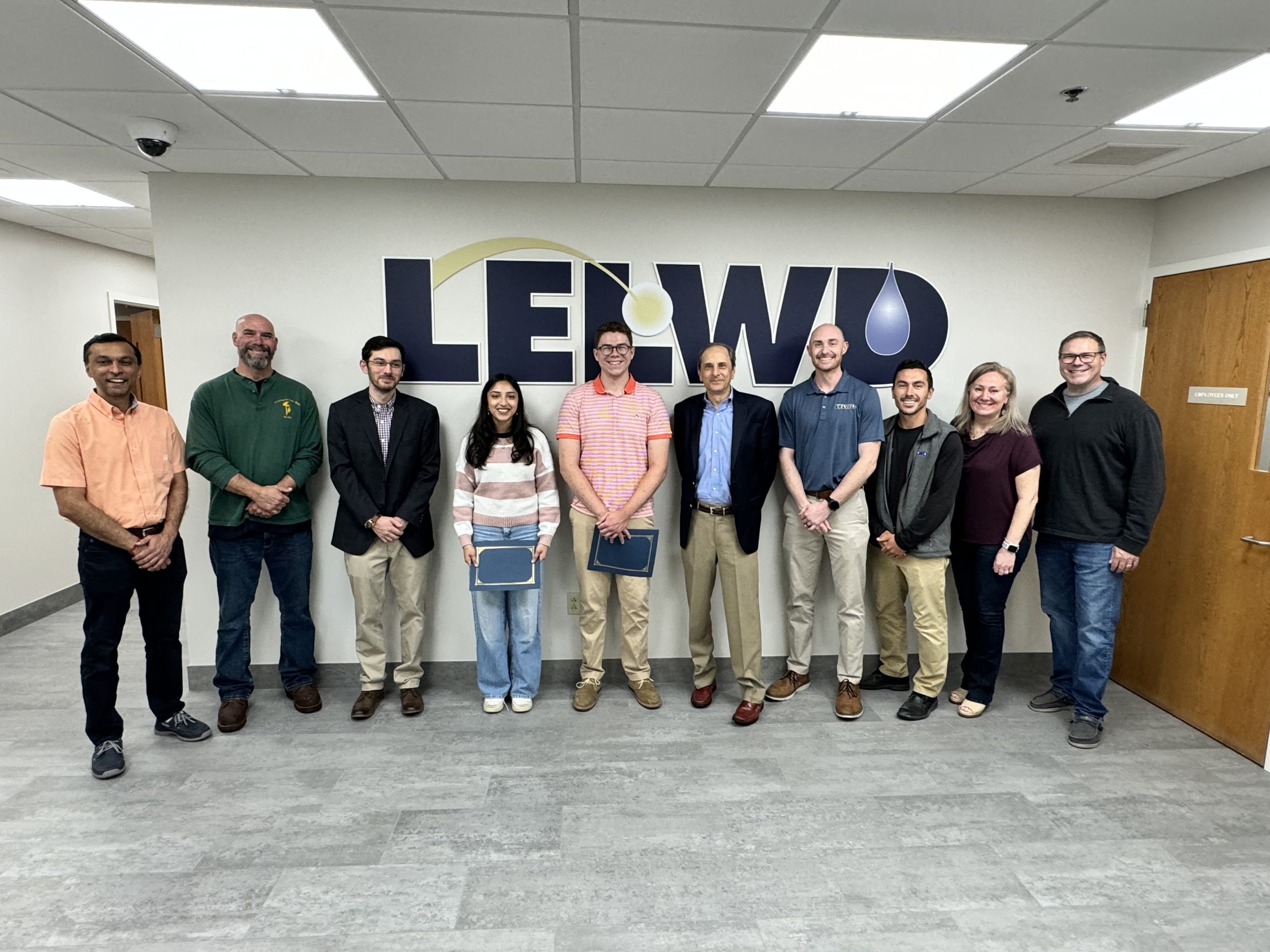 Eleven people pose for a group photo indoors in front of a wall bearing an LELWD logo.