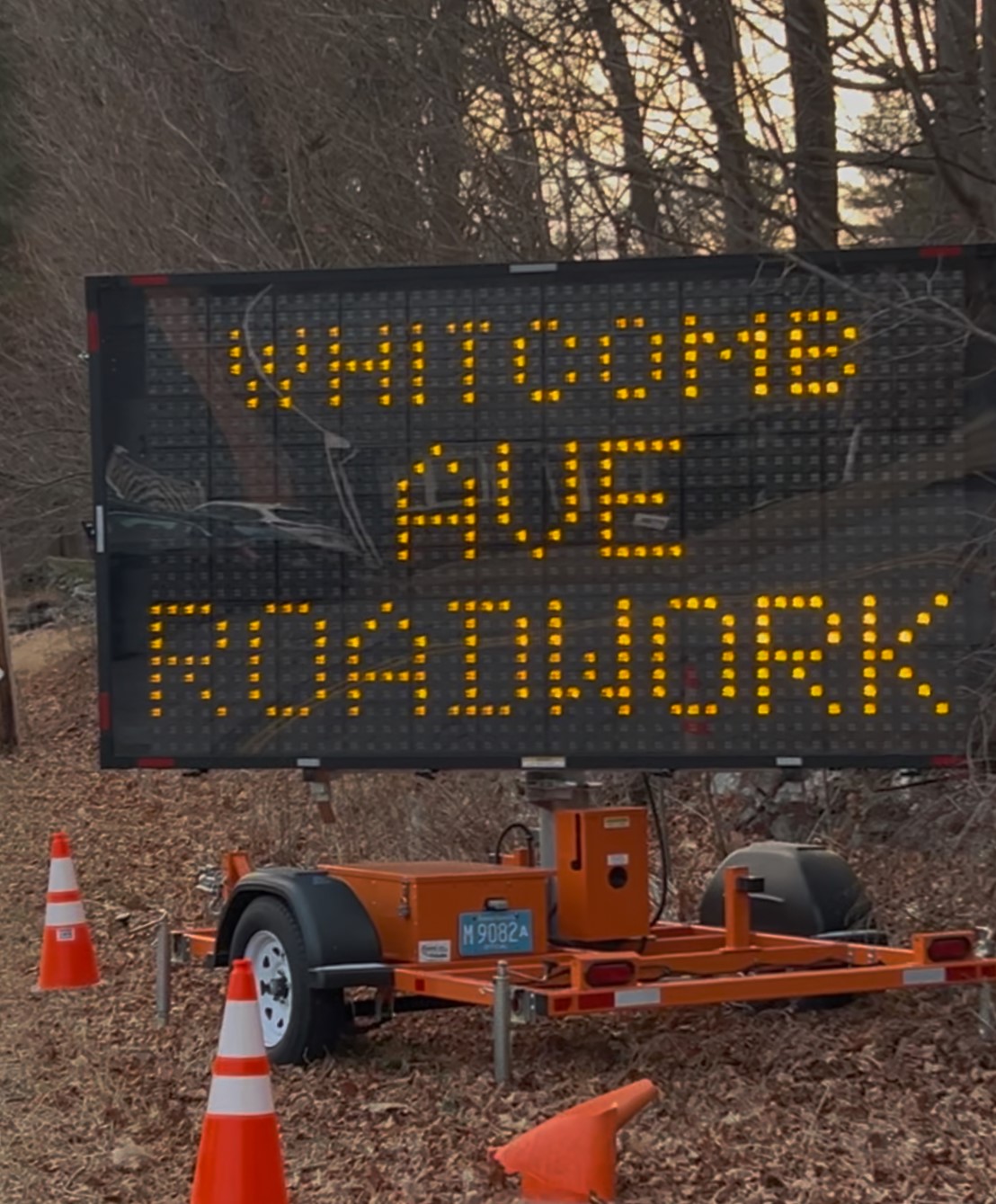 Portable electronic roadwork sign on a trailer reads WHITCOMBE AVE ROADWORK with orange traffic cones nearby.