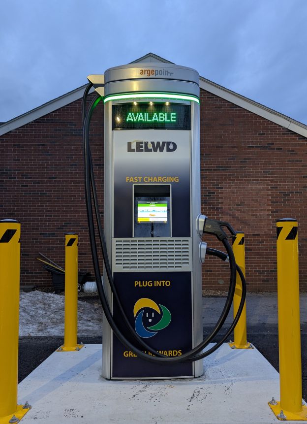 Electric vehicle charging station with an AVAILABLE sign, dual charging cables, and yellow bollards in front of a brick building.