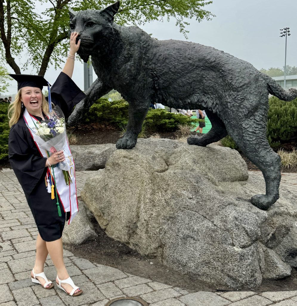 Cheerful graduate in cap and gown posing with a black wildcat statue perched on a rock.